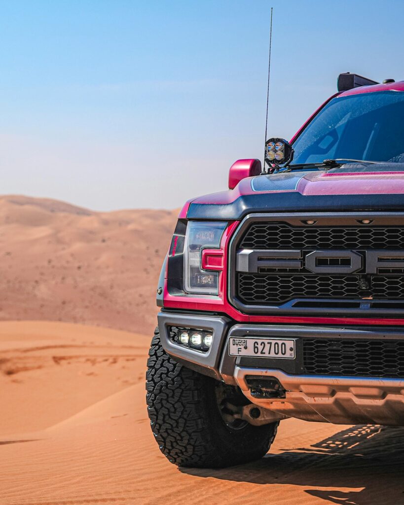 Red pickup truck on Dubai sand dunes under clear blue sky.