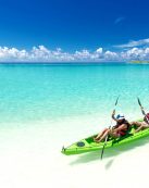 Two people kayaking in clear turquoise waters on a sunny tropical beach.