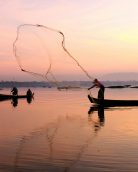 Silhouetted fishermen casting nets from boats during a serene sunrise over a calm lake.