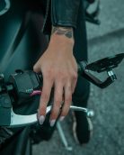 A detailed view of a hand gripping a motorcycle handlebar, showcasing tattoos and leather jacket.