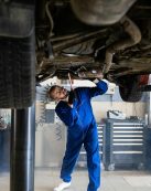 A female mechanic in blue coveralls inspects a car's undercarriage in an auto repair shop.
