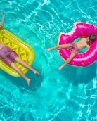 Two children lay on colorful floaties in a vibrant turquoise swimming pool, enjoying a summer day.